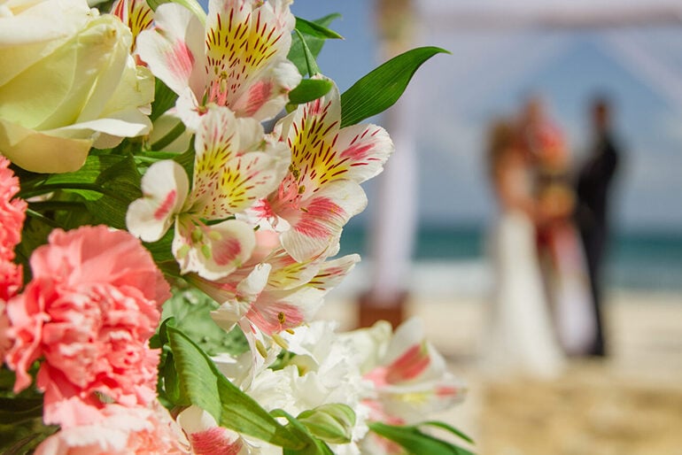 Bright pink tropical flowers in the foreground and a blurry hetero wedding couple on a beach in the background