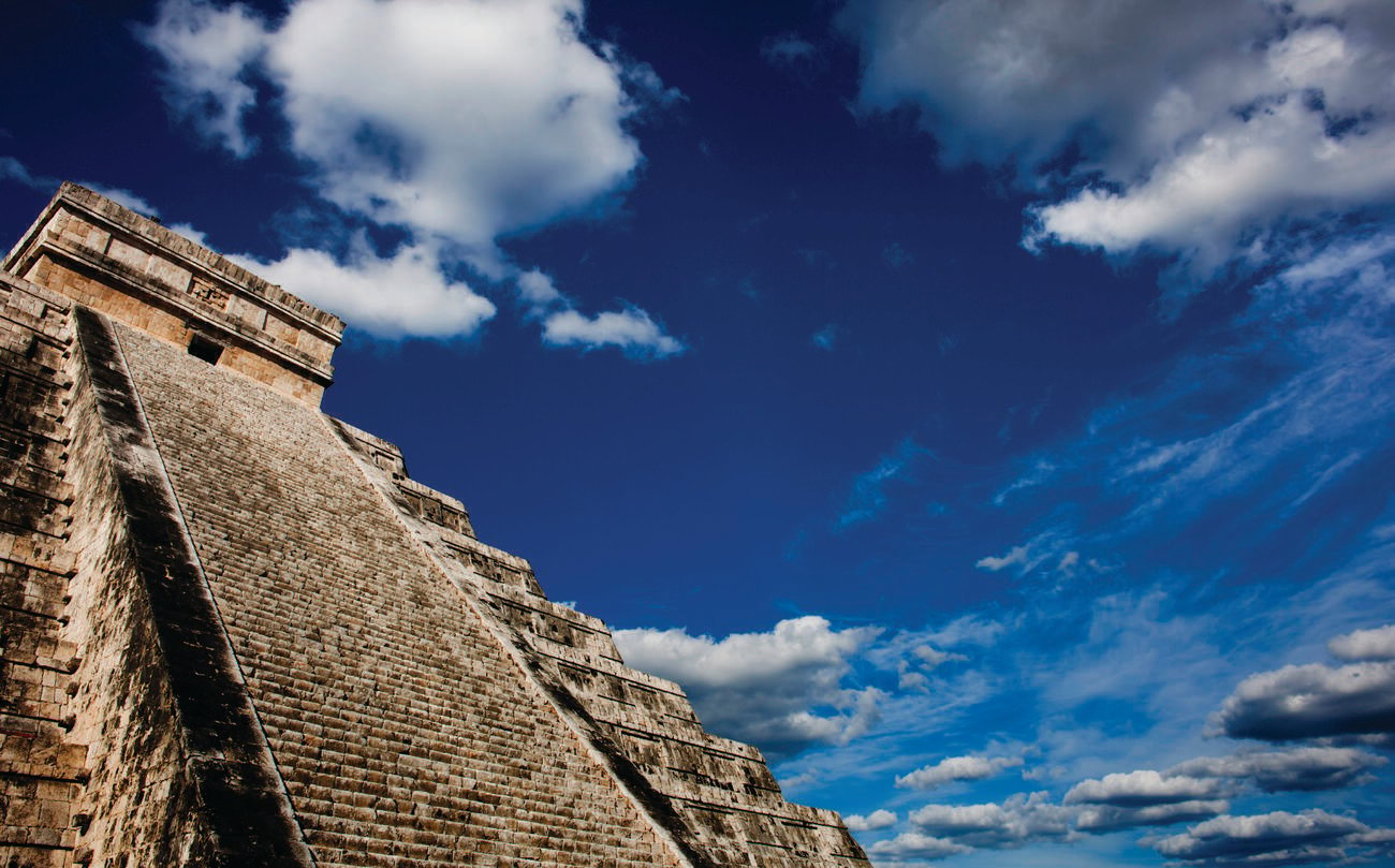 The ancient El Castillo pyramid at Chichen Itza against the backdrop of a bright blue sky with clouds.