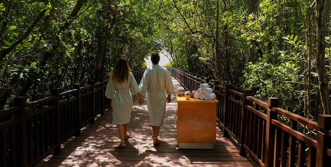 A couple walking on a jungle boardwalk with their faces turned away from the camera and a Champagne cart