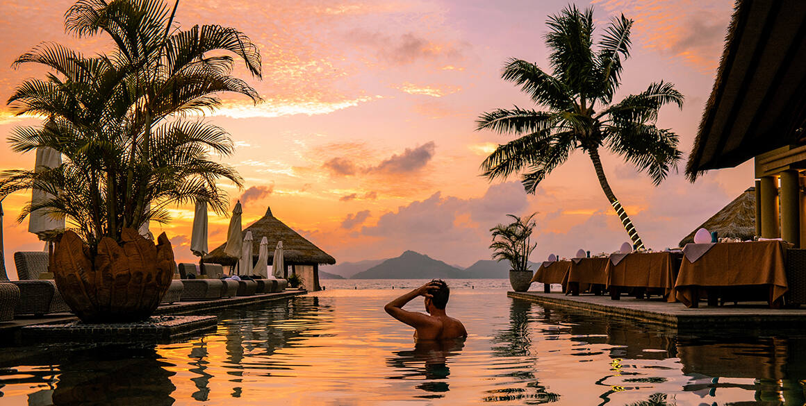 A pastel orange and pink sunset over an infinity-edge pool with one person's back to the camera