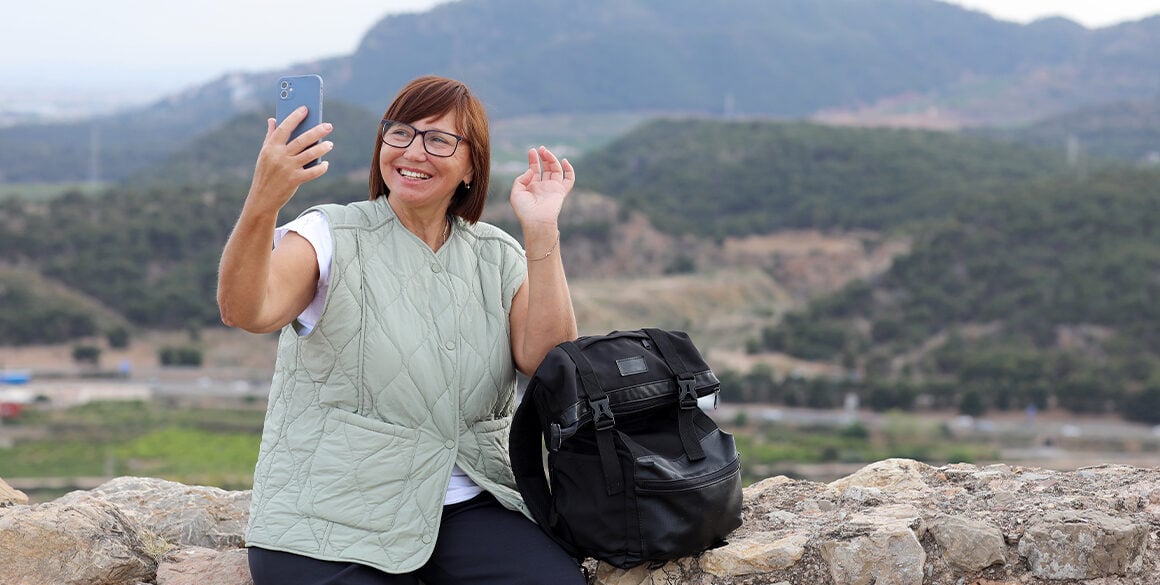 Middle-aged woman with a backpack to the side of her takes a photo with mountains in the background