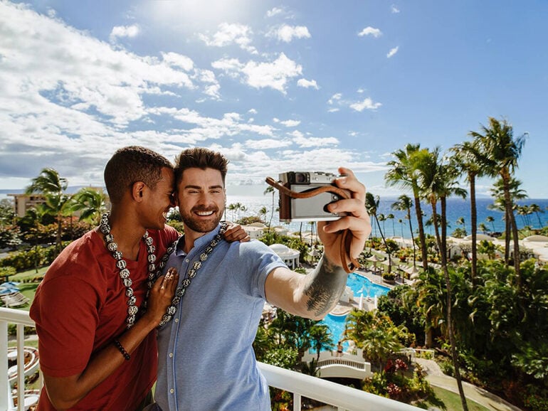 A Black and white man wearing leis embracing while taking a selfie on a balcony in Hawaii