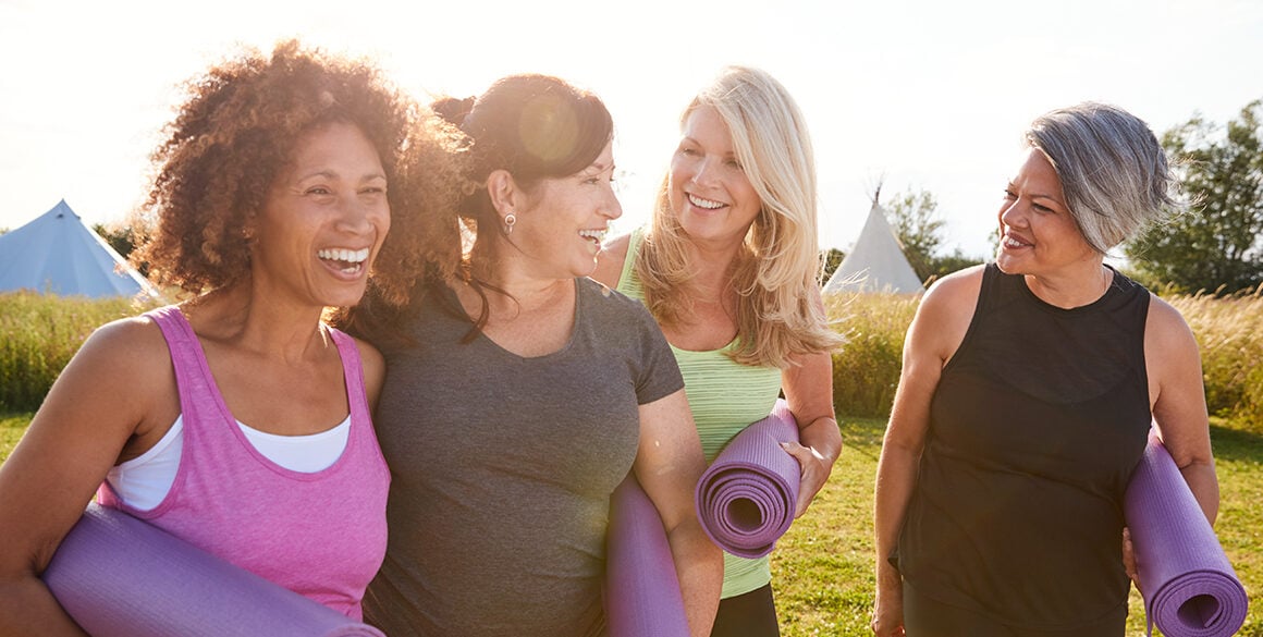 Four middle-aged women laughing and holding yoga mats in a grassy field