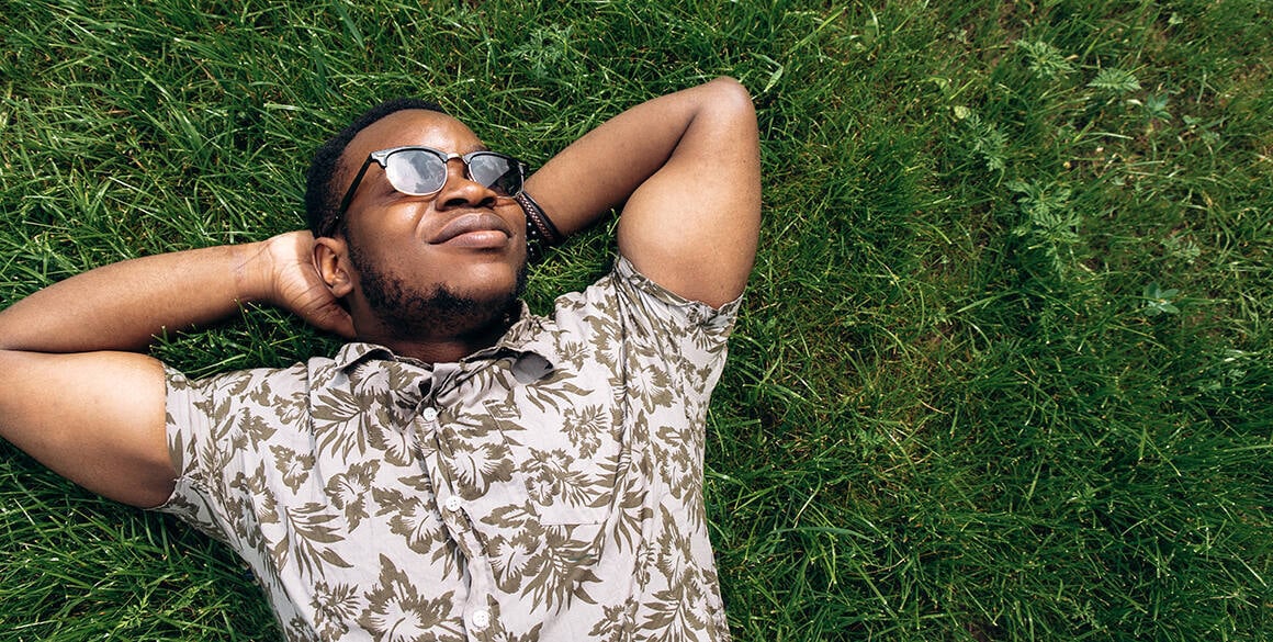 Black man with glasses in a casual button-down shirt lying down on grass, relaxing