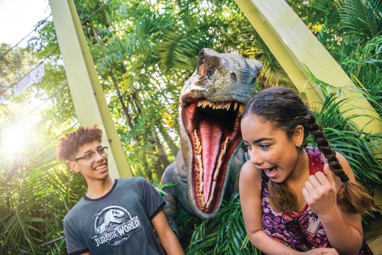 An animatronic velociraptor with its jaws wide open pops up between a laughing boy in a "Jurassic World" t-shirt and a screaming girl at Universal's Islands of Adventure theme park. 